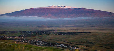 The beautiful and nearly 14,000 foot tall Hawaiian Volcano?Mauna Kea?looms over the landscape on the Big Island of Hawaii. Rolling hills in the foreground give way to vast countryside. Hawaii's tallest volcano is snow capped and rises well above the thin cloud layer.